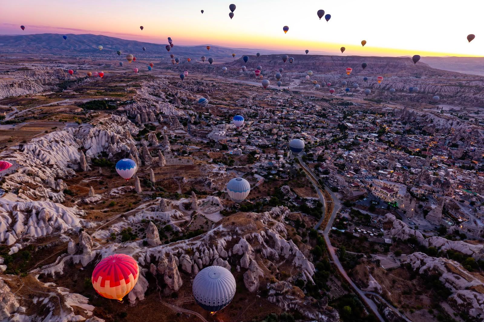 Numerous hot air balloons float over a rocky valley and town at sunrise.