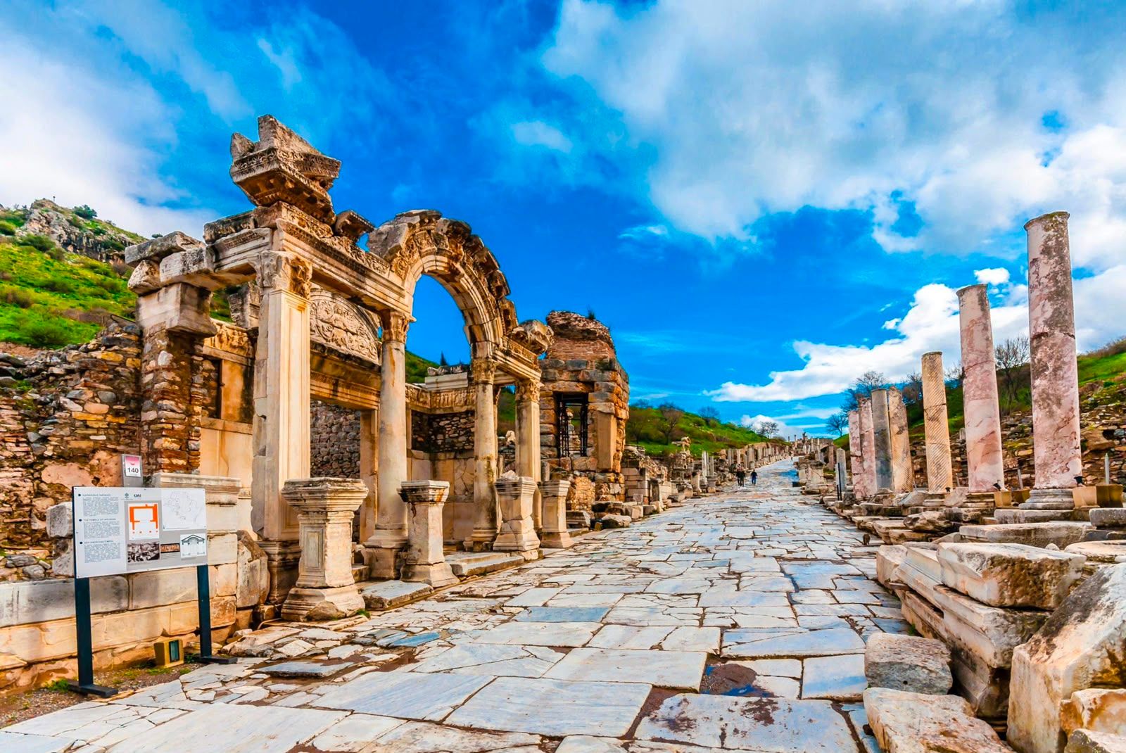 An ancient stone road runs past ruined marble columns under a blue sky.