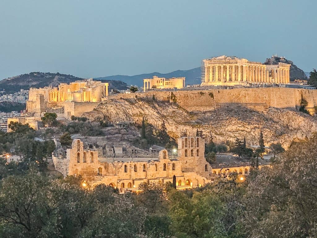 Illuminated Acropolis of Athens, featuring the Parthenon and Odeon, at dusk.