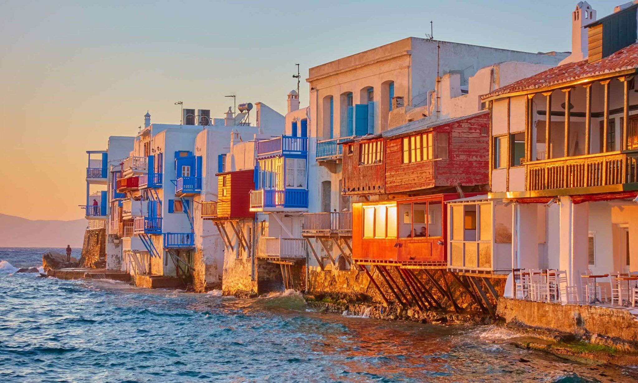 Whitewashed houses with colorful balconies extending over the Aegean Sea at sunset.