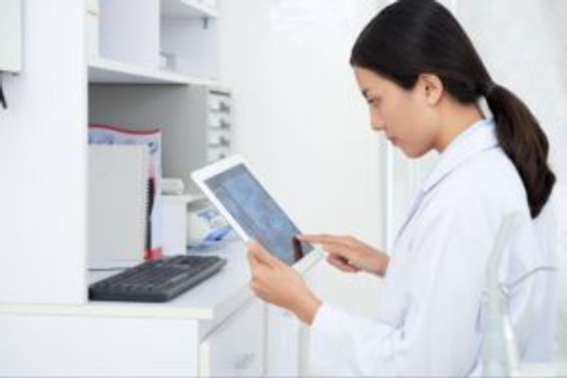 A woman in a white lab coat uses a tablet while sitting at a desk with a computer keyboard, notebooks, and shelves in a bright office or laboratory setting.