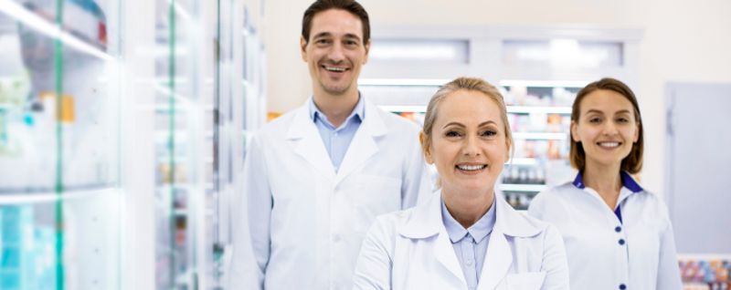 Three smiling healthcare professionals, two women and one man, stand in a brightly lit pharmacy. They are wearing white lab coats, suggesting they are pharmacists or pharmaceutical staff. Shelves filled with products are visible in the background.