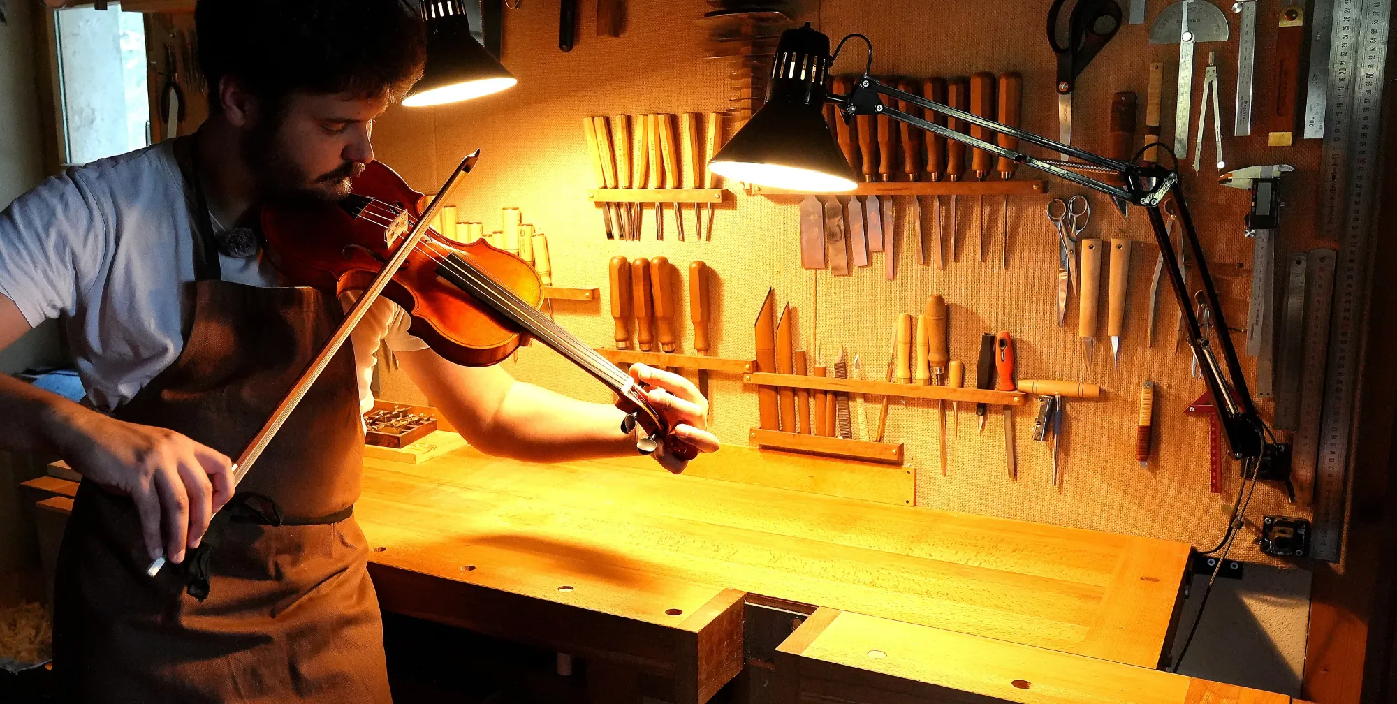Nicolas Wicky joue un violon debout devant son établi, sous les lampes de l'atelier.