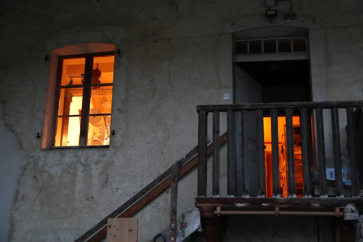 La façade de l'atelier : une fenêtre éclairée où l'on voit un violon suspendu, et une porte ouverte en haut d'un escalier en bois.