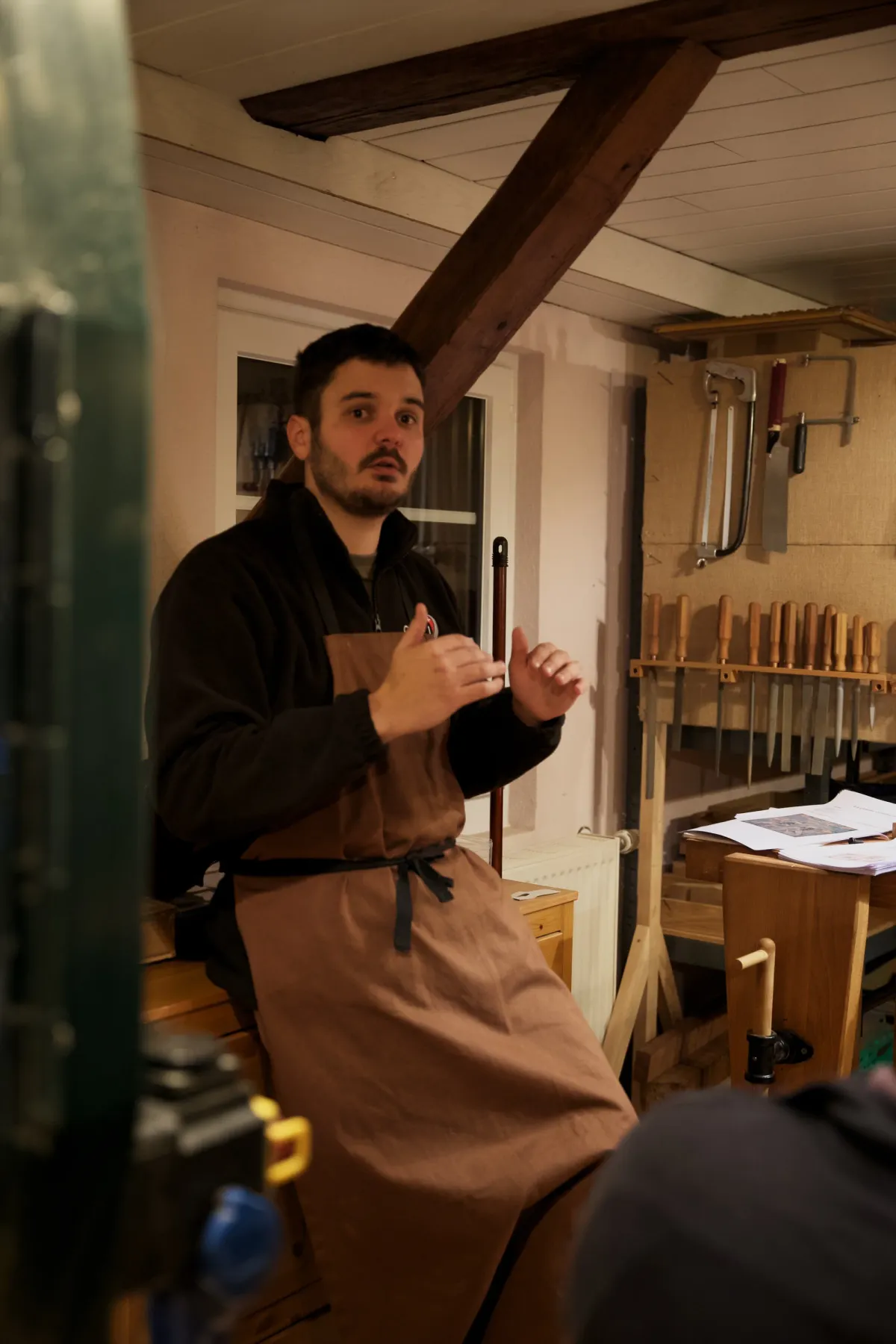 Nicolas Wicky debout dans son atelier, en tablier, expliquant un geste devant le mur d'outils.