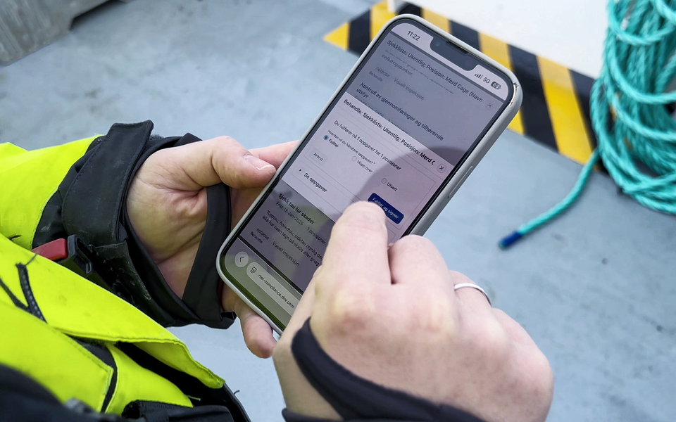 Person i arbeidsklær holder en mobiltelefon og registrerer informasjon i en app, stående i et industrielt miljø med tauverk og gulvmerking i bakgrunnen.