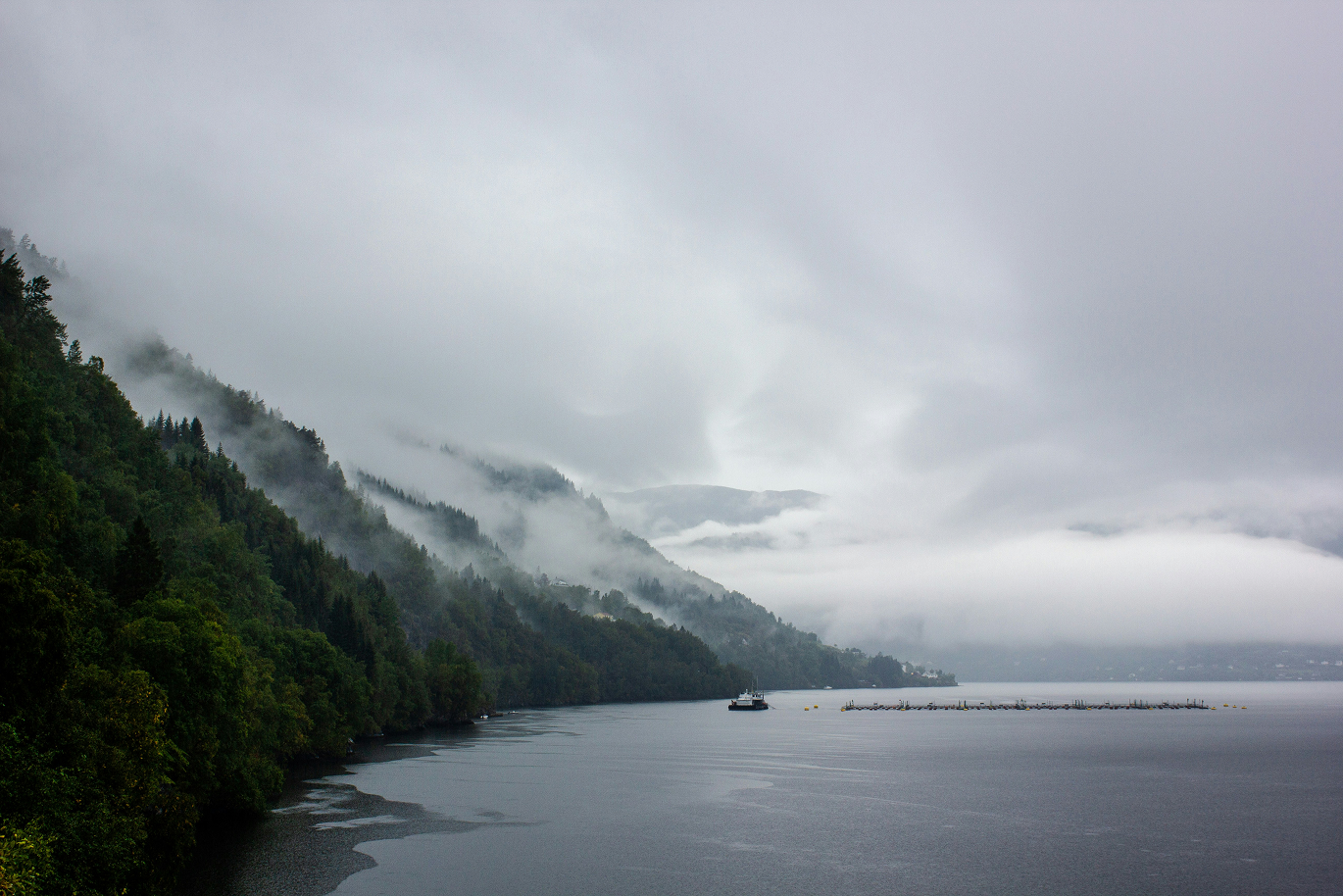 Tåkelagt fjordlandskap med skogkledde fjellsider som stuper ned i stille vann, der et lite fartøy og et oppdrettsanlegg ligger delvis skjult i disen.
