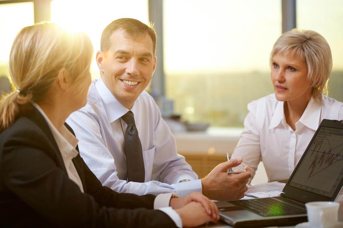 A man and two women are sitting at a table discussing over a laptop.