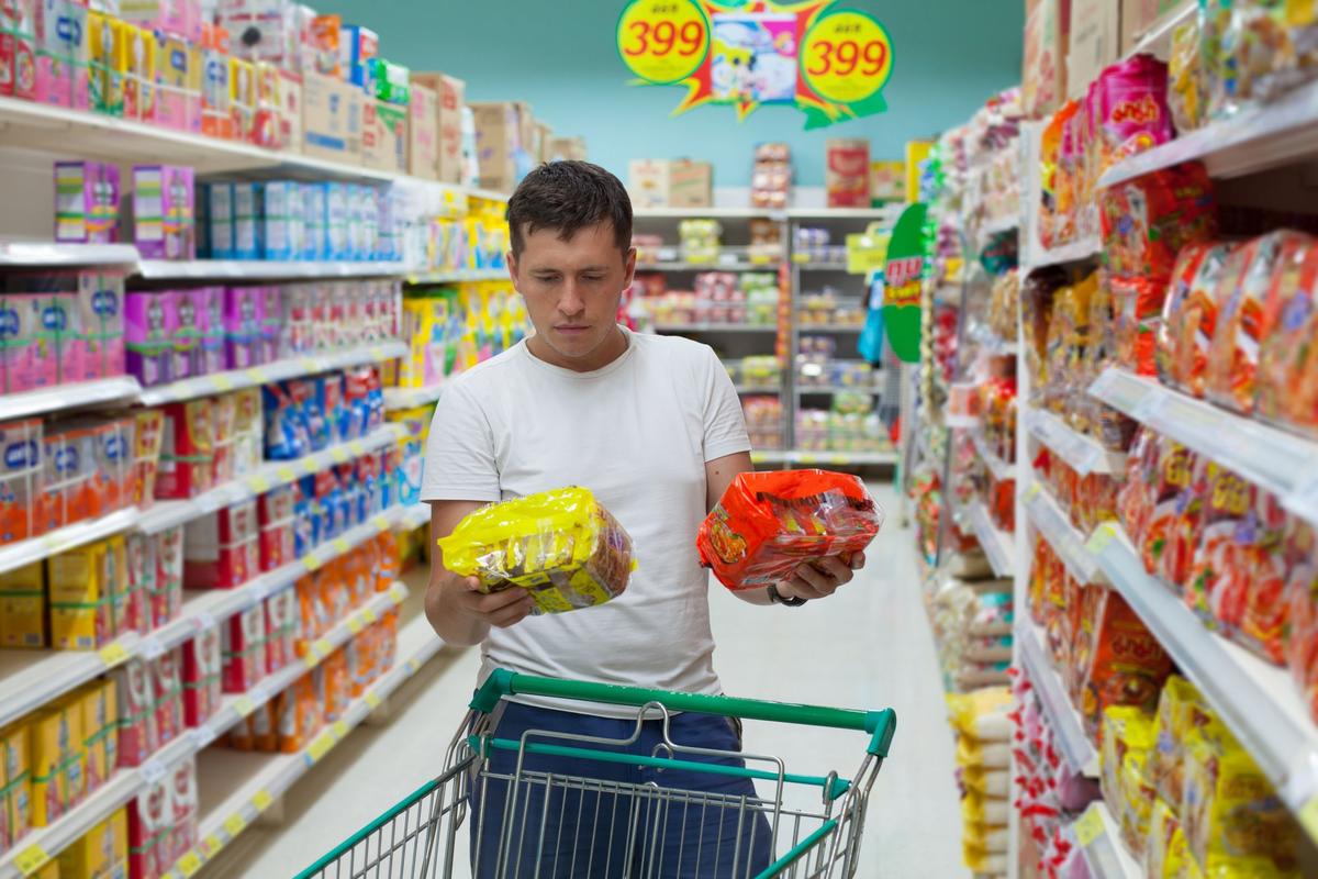A man in a white shirt carrying a cart loaded with groceries is holding two items, wondering which one he should get. 