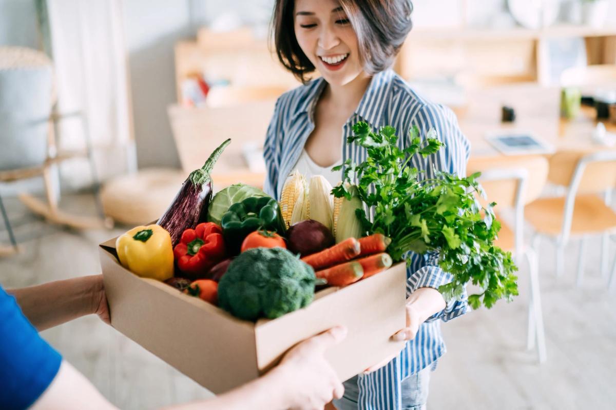 A man is handing a box of vegetables to the woman in a striped blue top.