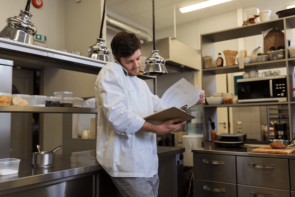 A man in a white coat holding a clipboard and talking on the phone in the kitchen.