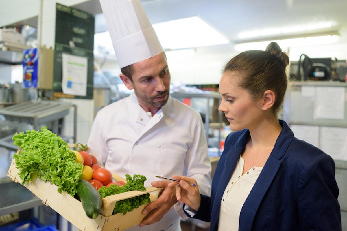 A chef wearing a chef hat holding a crate of vegetables, talking to a woman.