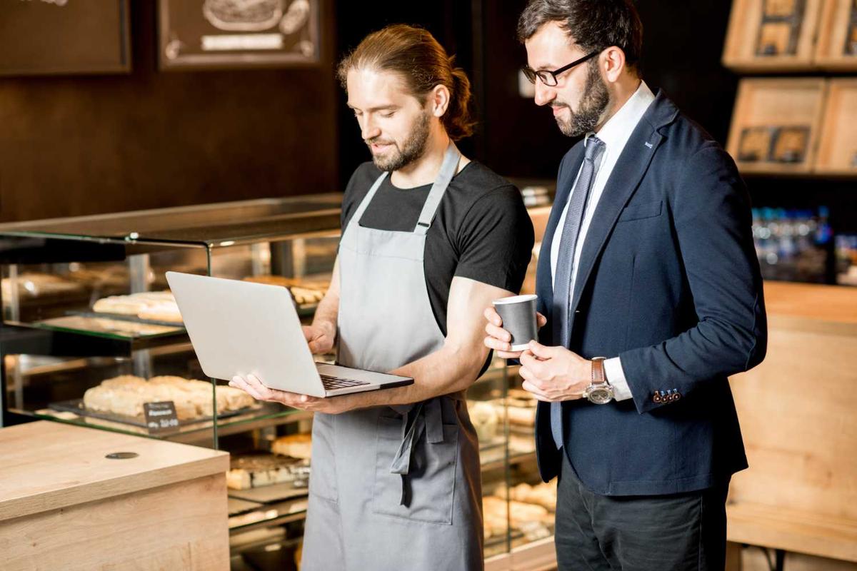 A man wearing an apron is holding a laptop, and the other man is wearing a suit while holding a coffee.