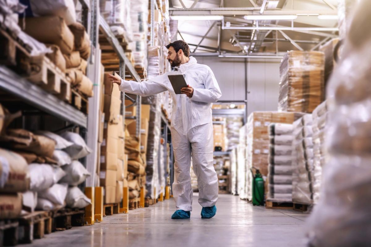 A man in white jumpsuit holding a tablet while carefully checking the stocks in a warehouse.