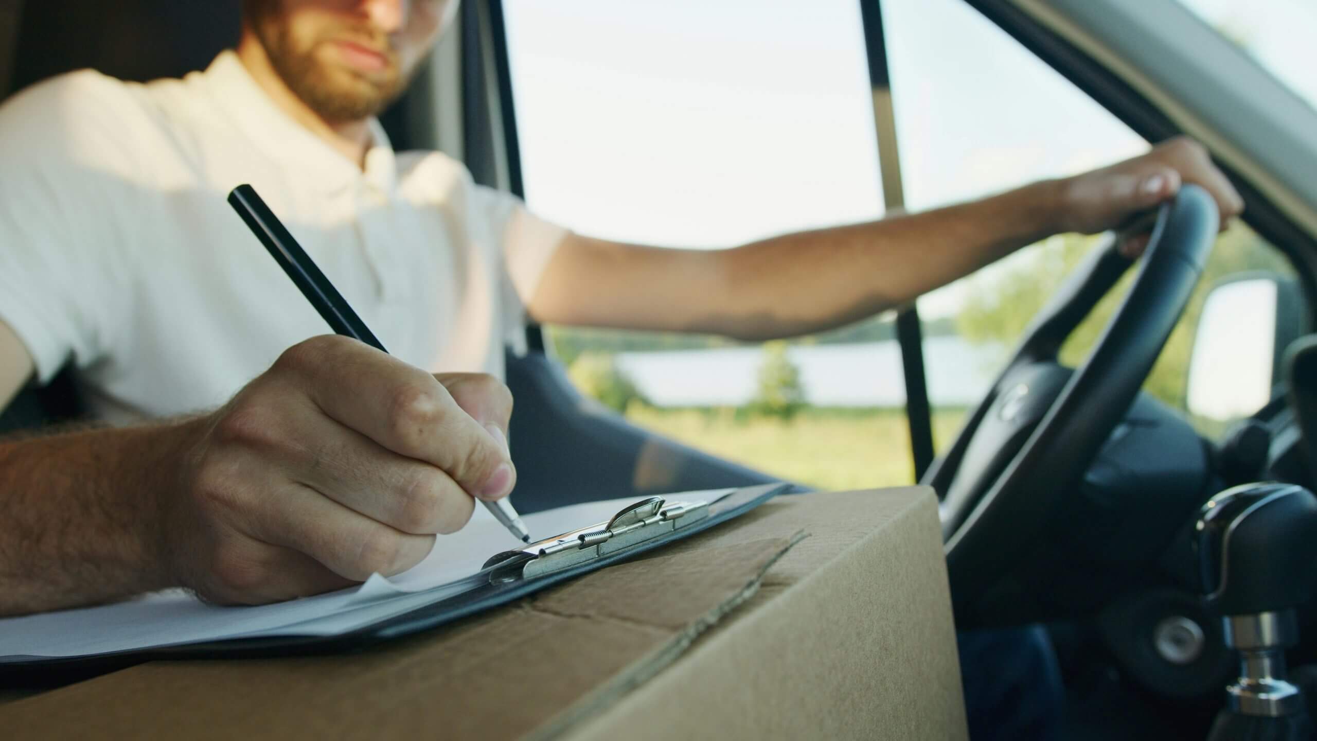 Delivery driver in a van signing paperwork on a clipboard over a cardboard box.