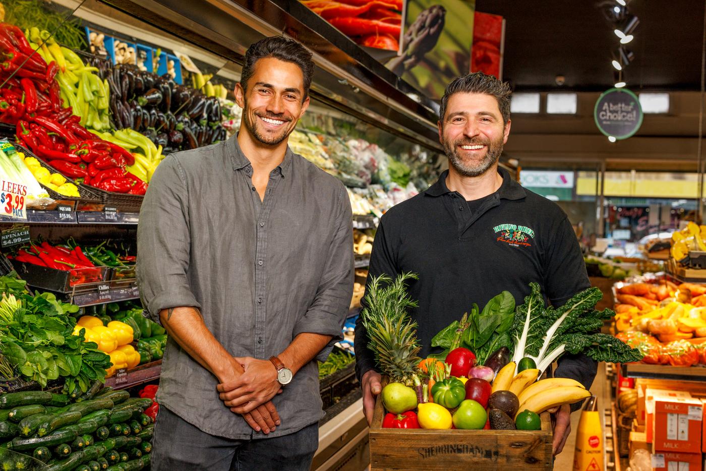 Two men standing in front of a produce display holding a crate of vegetables.