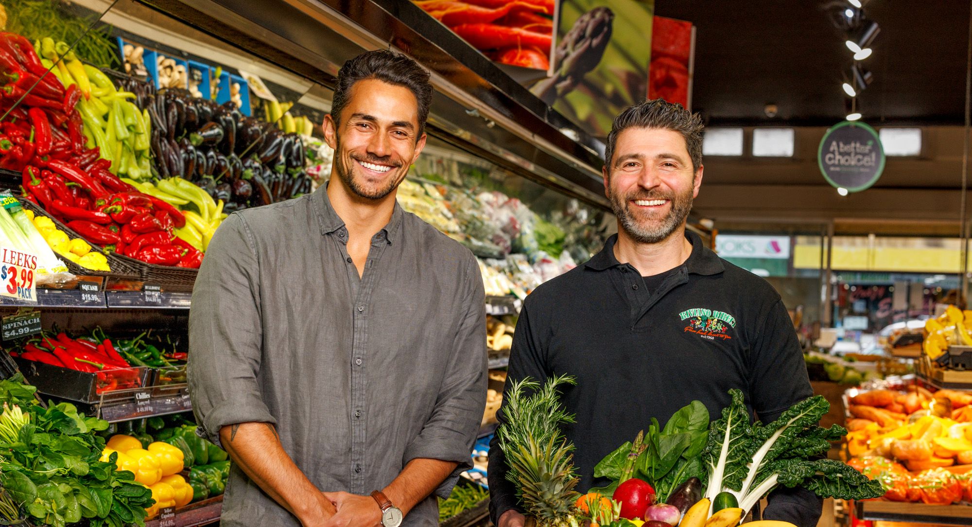 Two men standing in front of a produce display holding a crate of vegetables.