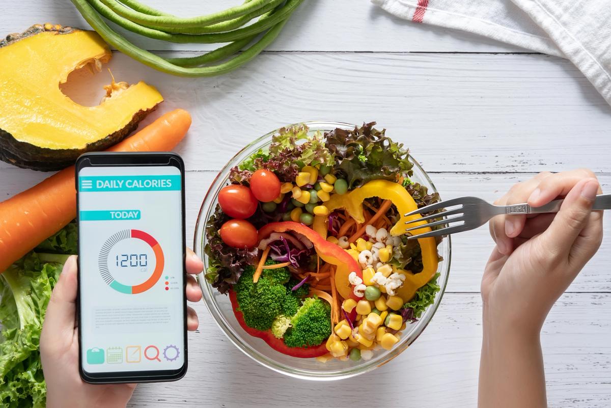 A close-up of a diet app on a mobile device, a hand holding a fork, and a bowl of fresh fresh fruits and vegetables on a wooden table.