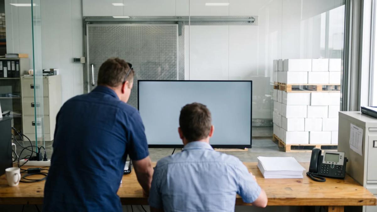 Two order desk staff at a food wholesaler sharing one large monitor while a stack of printed order sheets sits beside them.