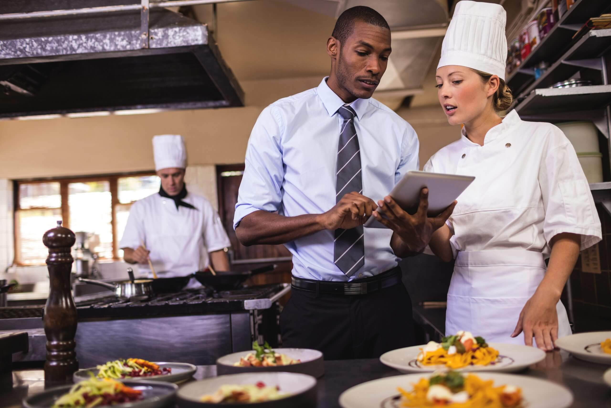 A chef holding a tablet, discussing with the other chef in the kitchen.
