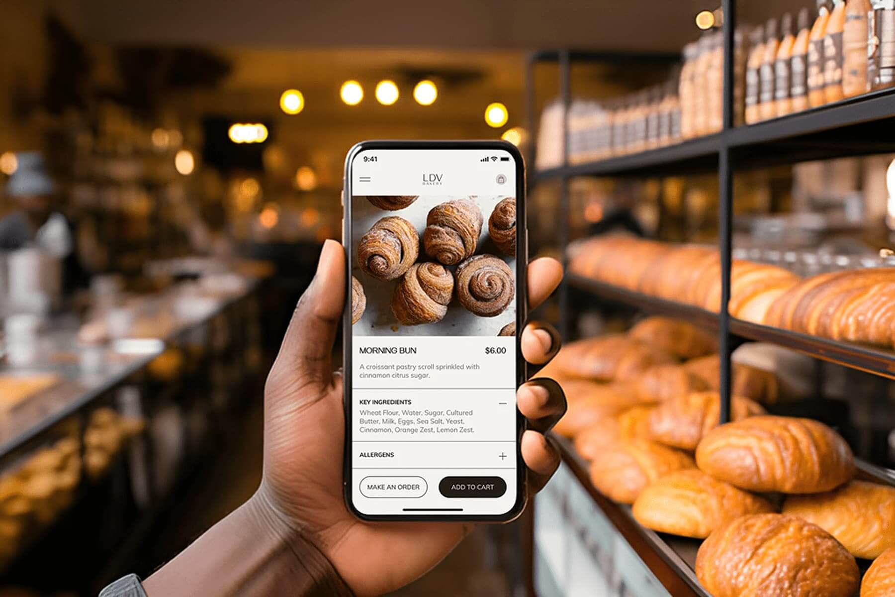 Hand holding a phone showing a bakery ordering app in front of shelves of fresh bread.