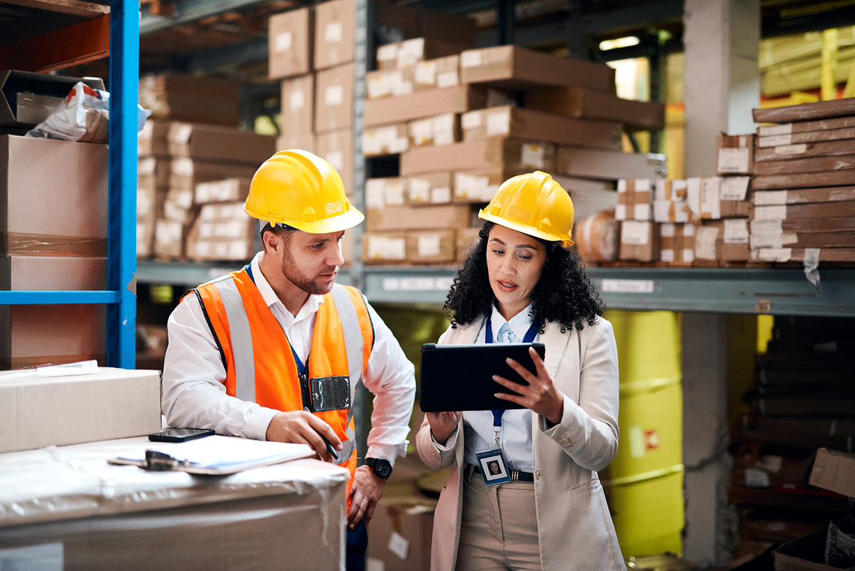 A man and woman wearing hard hats looking at a tablet in a warehouse.
