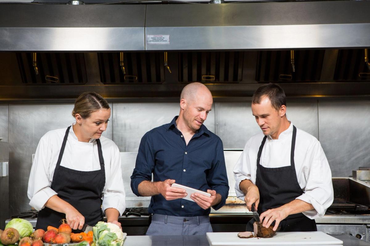 Two kitchen staff are preparing food, and a man between them is holding a tablet.