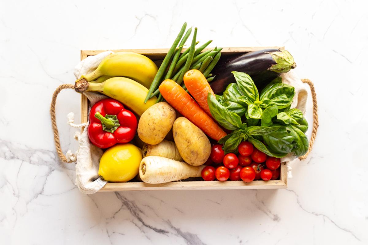 A wooden box filled with fresh vegetables and fruits, displayed on a white marble countertop, showcasing vibrant colors.
