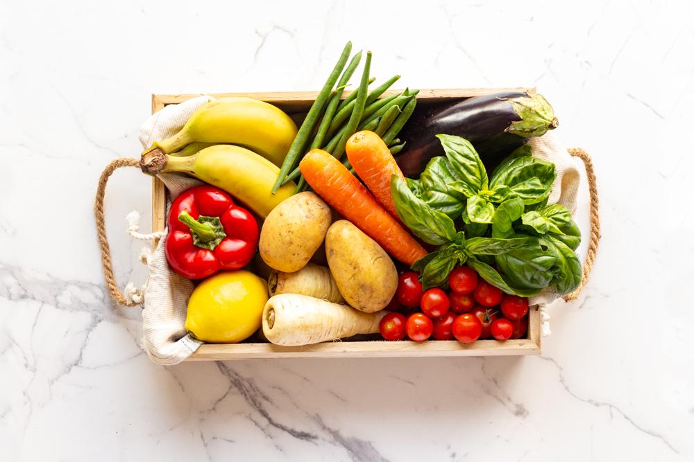 A wooden box filled with fresh vegetables and fruits, displayed on a white marble countertop, showcasing vibrant colors.