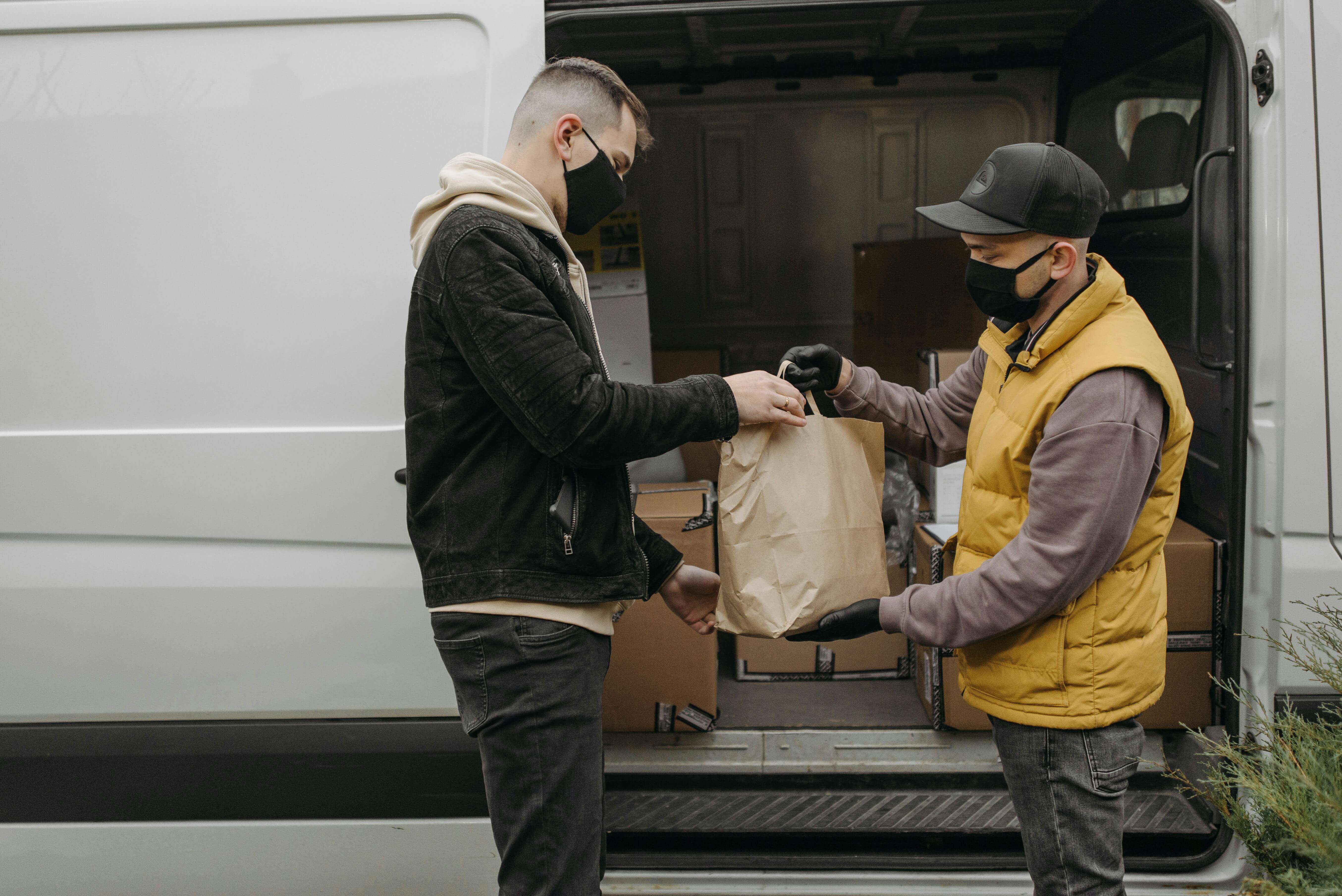 Two masked people exchanging a paper bag from the back of a delivery van.