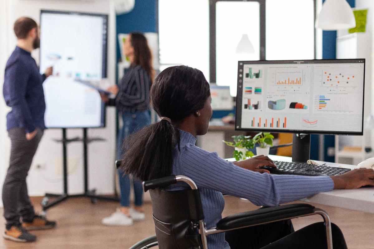 A woman in a wheelchair is using a computer with a pricing graph shown on the screen.
