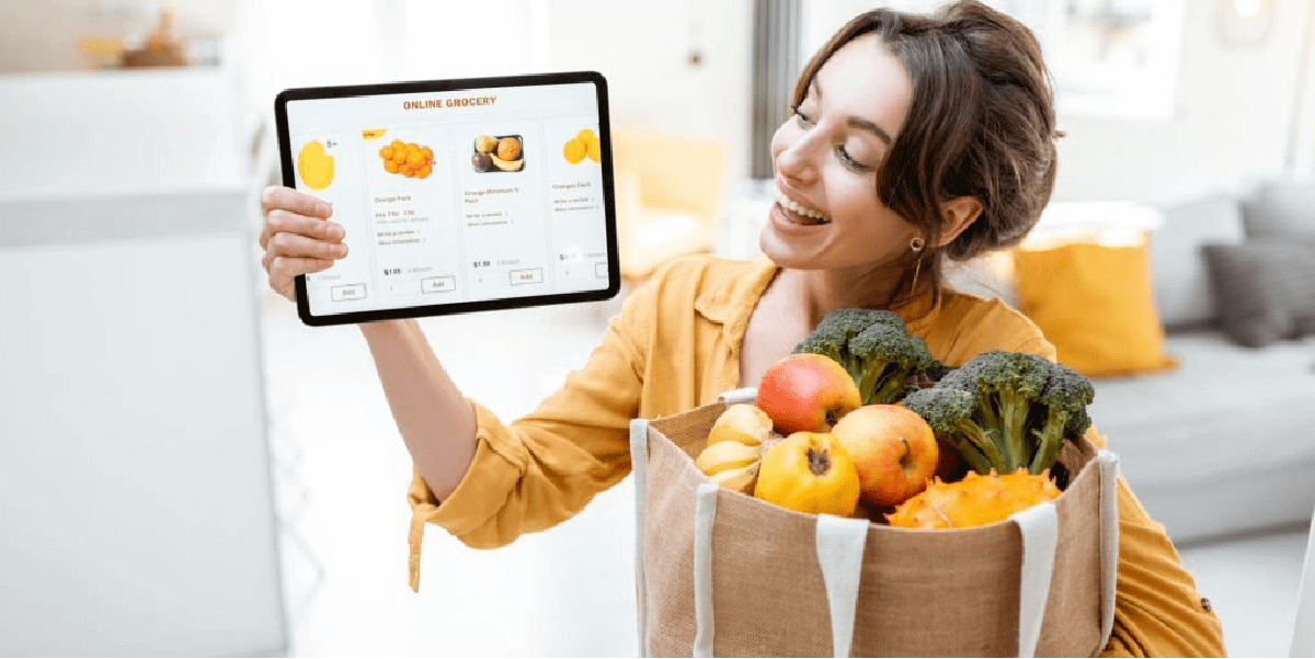Happy woman holding a tablet with a grocery app on screen and a bag of groceries in her other hand.