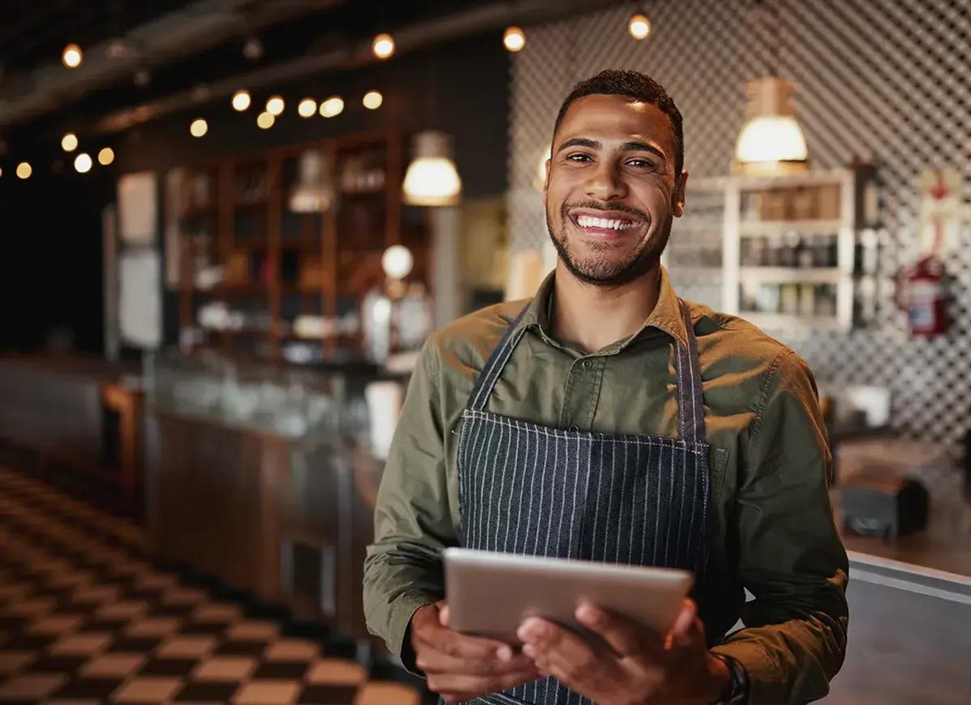 A barista in a modern cafe holding a tablet, standing and smiling confidently with warm lighting and blurred customers in the background.