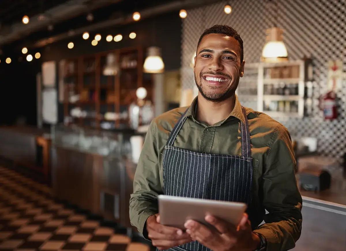 A barista in a modern cafe holding a tablet, standing and smiling confidently with warm lighting and blurred customers in the background.
