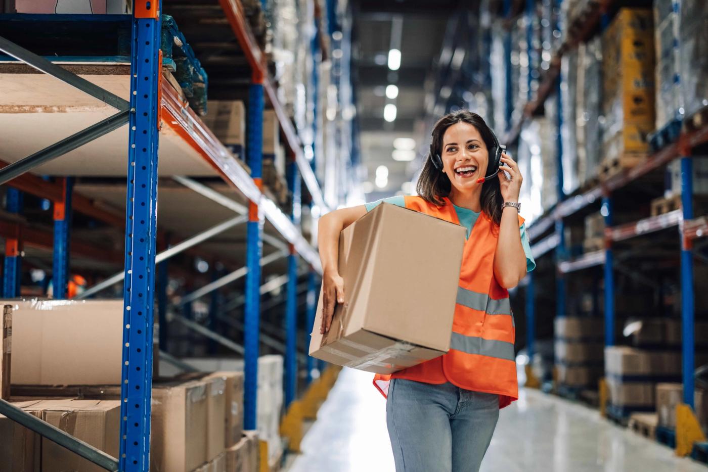 A woman standing inside a warehouse, holding a box while talking on the phone.