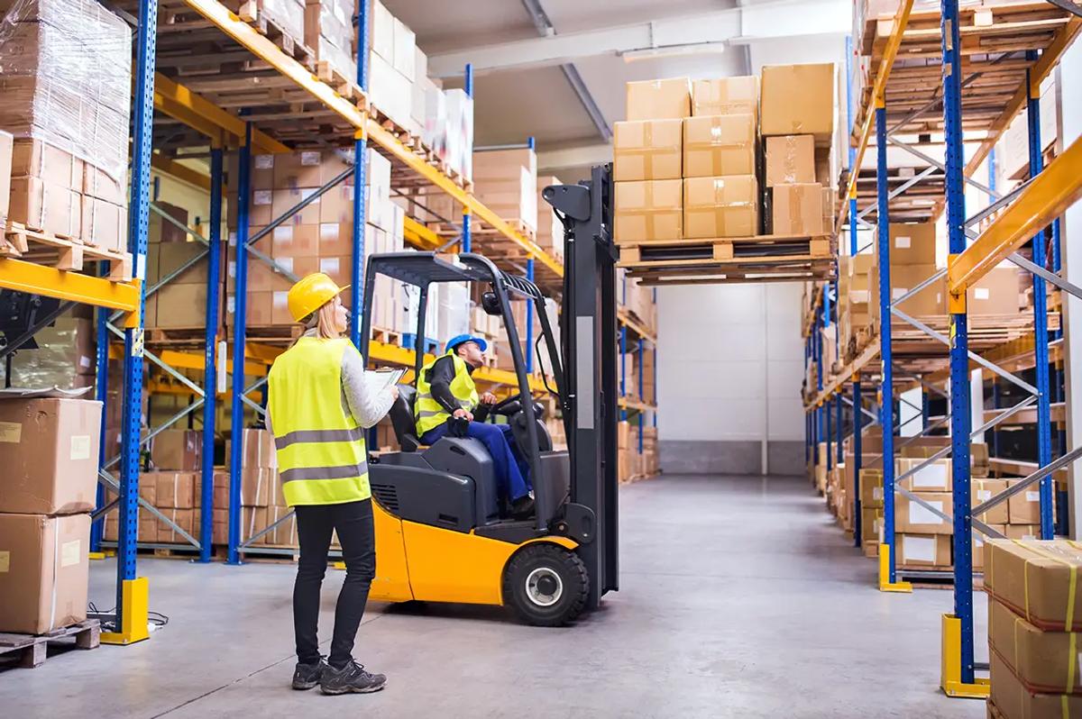 Workers in safety vests and helmets operate a forklift in a warehouse filled with stacked boxes and shelves.
