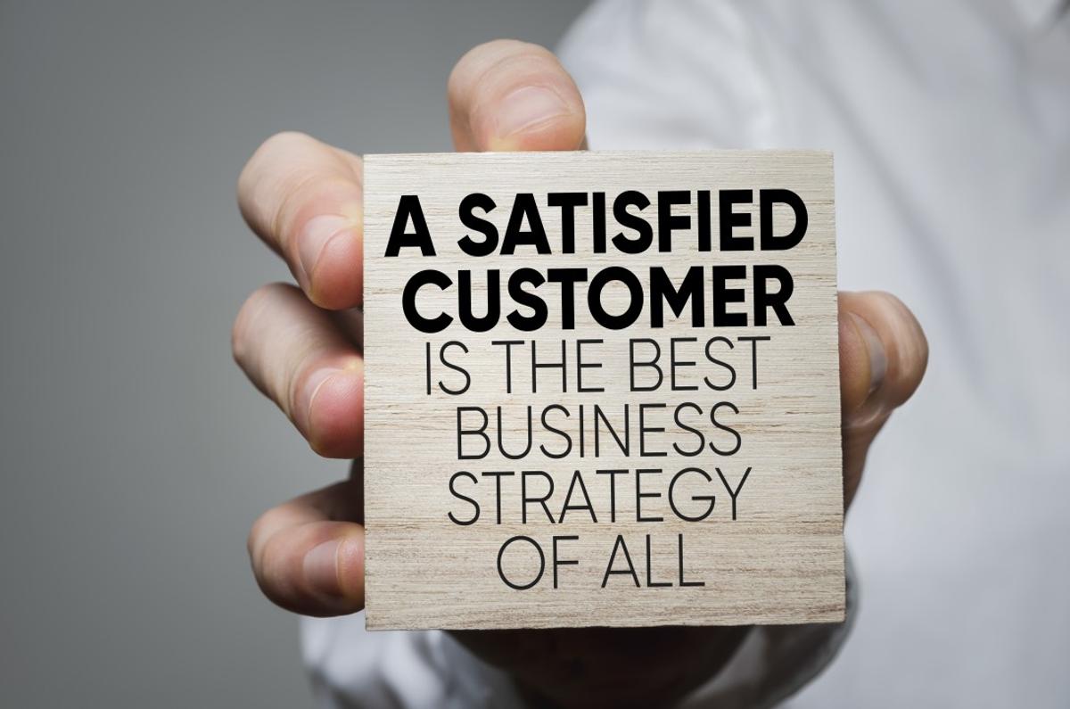 A hand holding a wooden board with the word "A satisfied customer is the best business strategy of all".