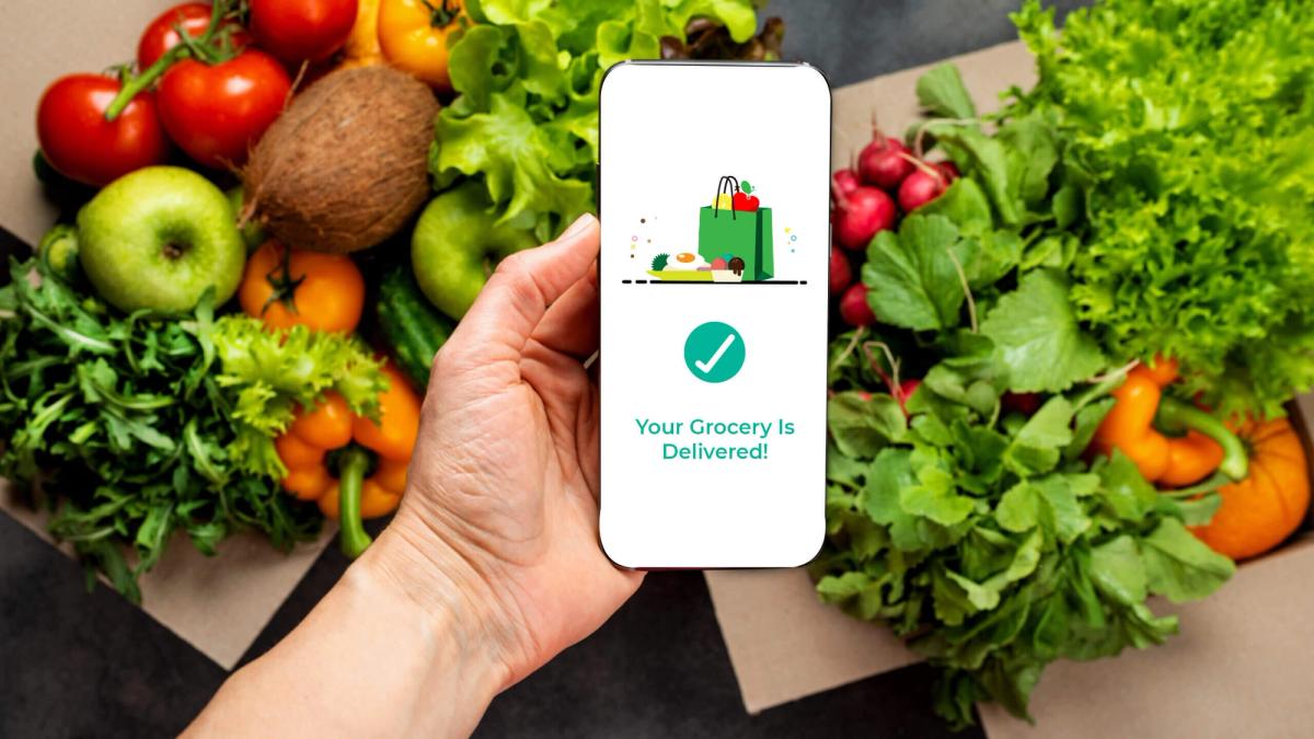 A hand holding a cellphone with fresh produce on the background showing a notification that grocery is delivered.