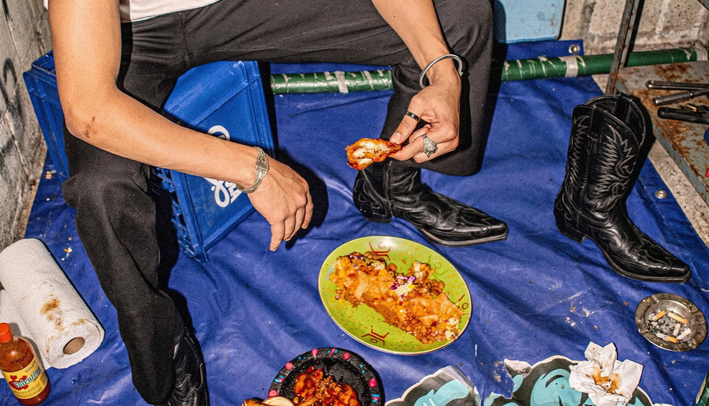 Comensal con botas vaqueras sentado en el suelo sobre una manta azul, junto a un plato de comida mexicana