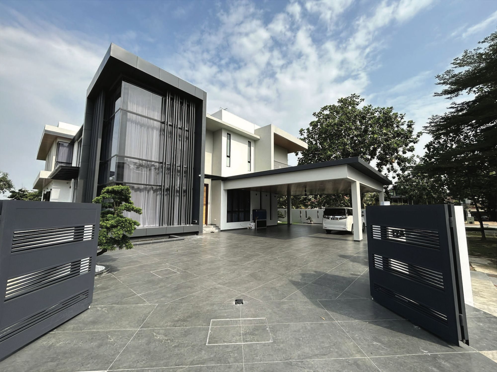 The 2-Storey Bungalow in Bukit Rimau seen through its open gates: white two-storey volumes, a dark double-height glazed bay with vertical fins and a flat-roofed car porch over a tiled forecourt