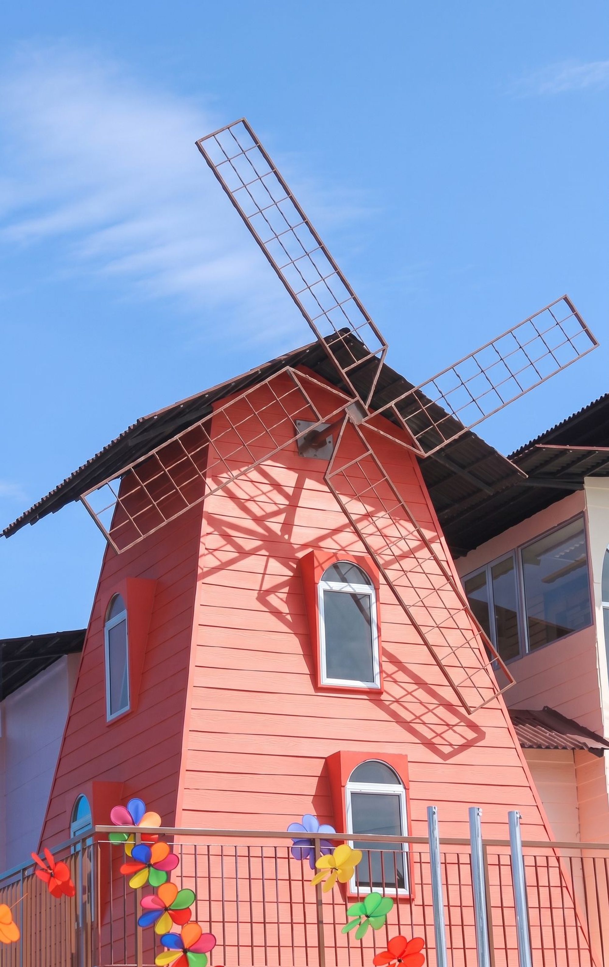 The red windmill tower against a blue sky, with small arched windows and colourful pinwheels on the fence