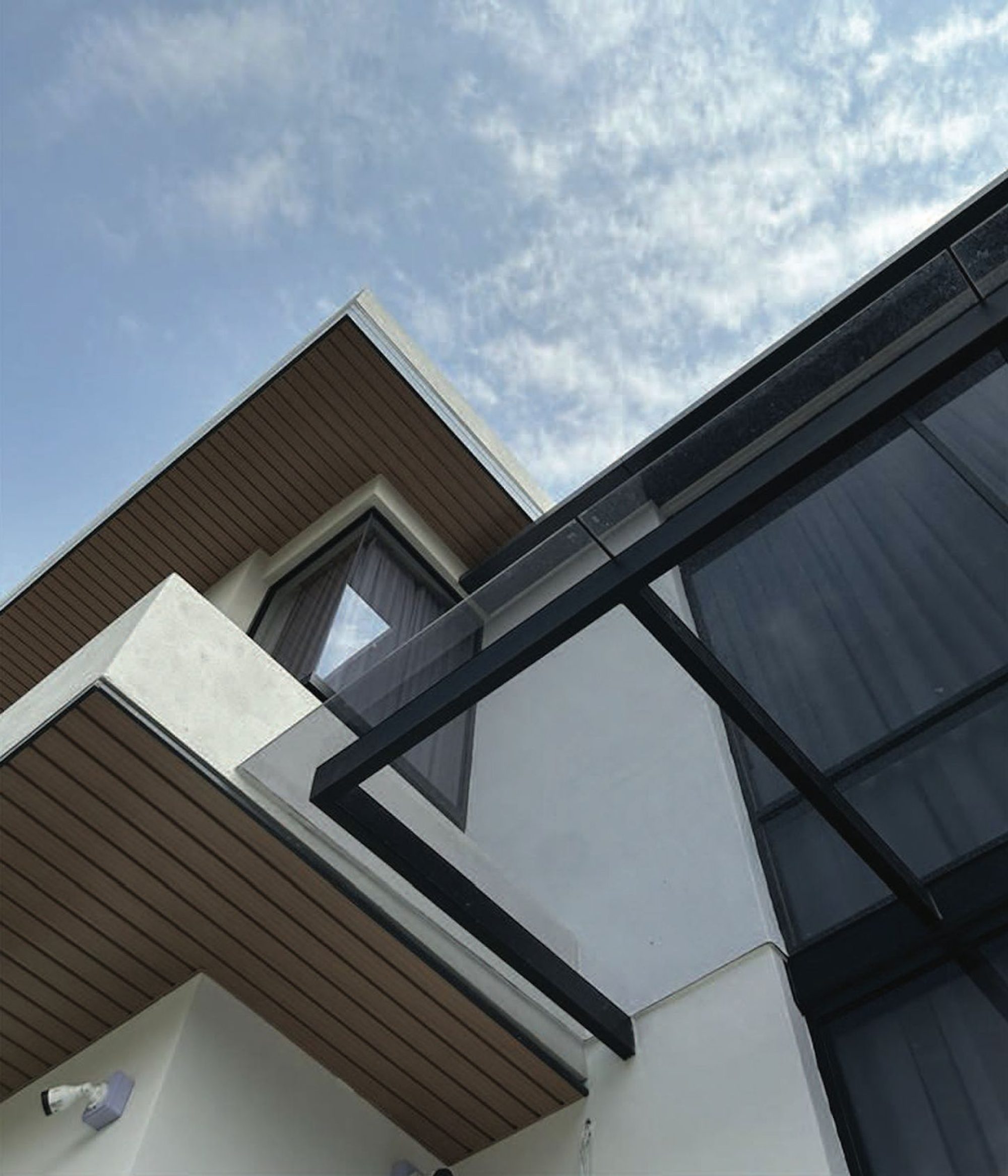 Looking up at white cantilevered eaves with wood-grain soffits and a black steel-framed glass canopy against the sky