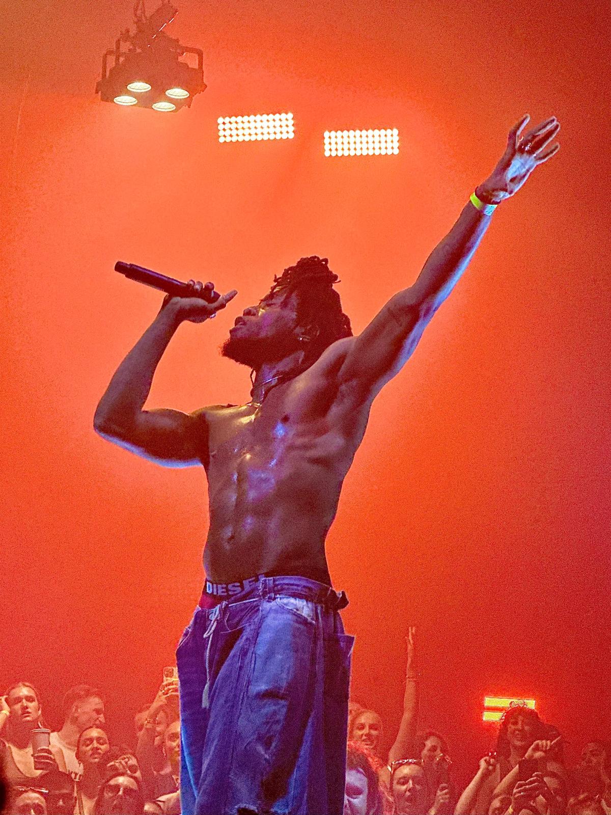 A shirtless singer performs on stage against a red backdrop.