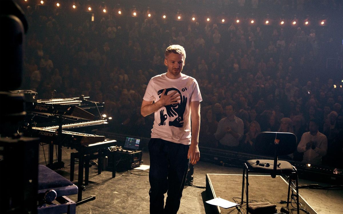 A bearded man in a white t-shirt stands onstage at a concert and looks pleased.