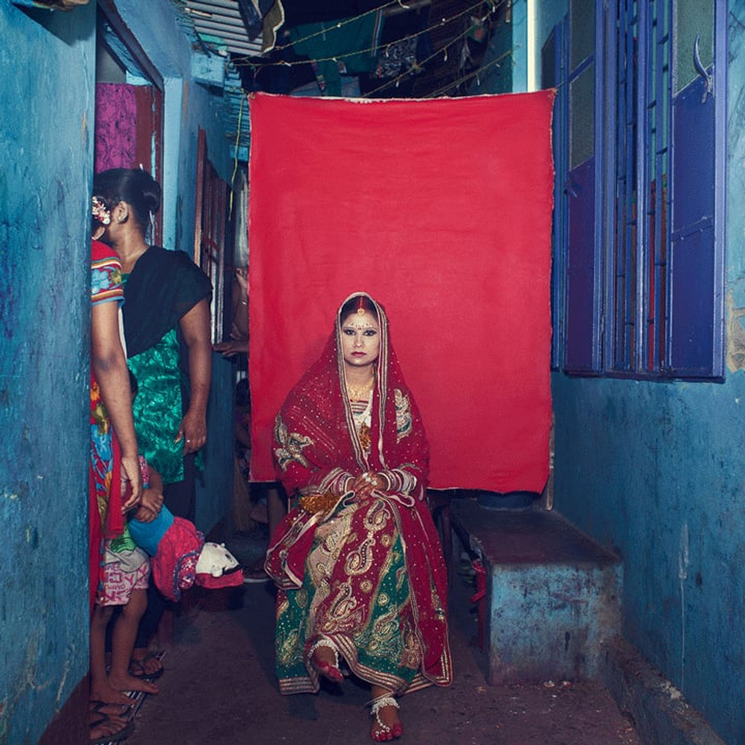 Person in red-and-gold ceremonial clothing sits while onlookers gather at the edges.
