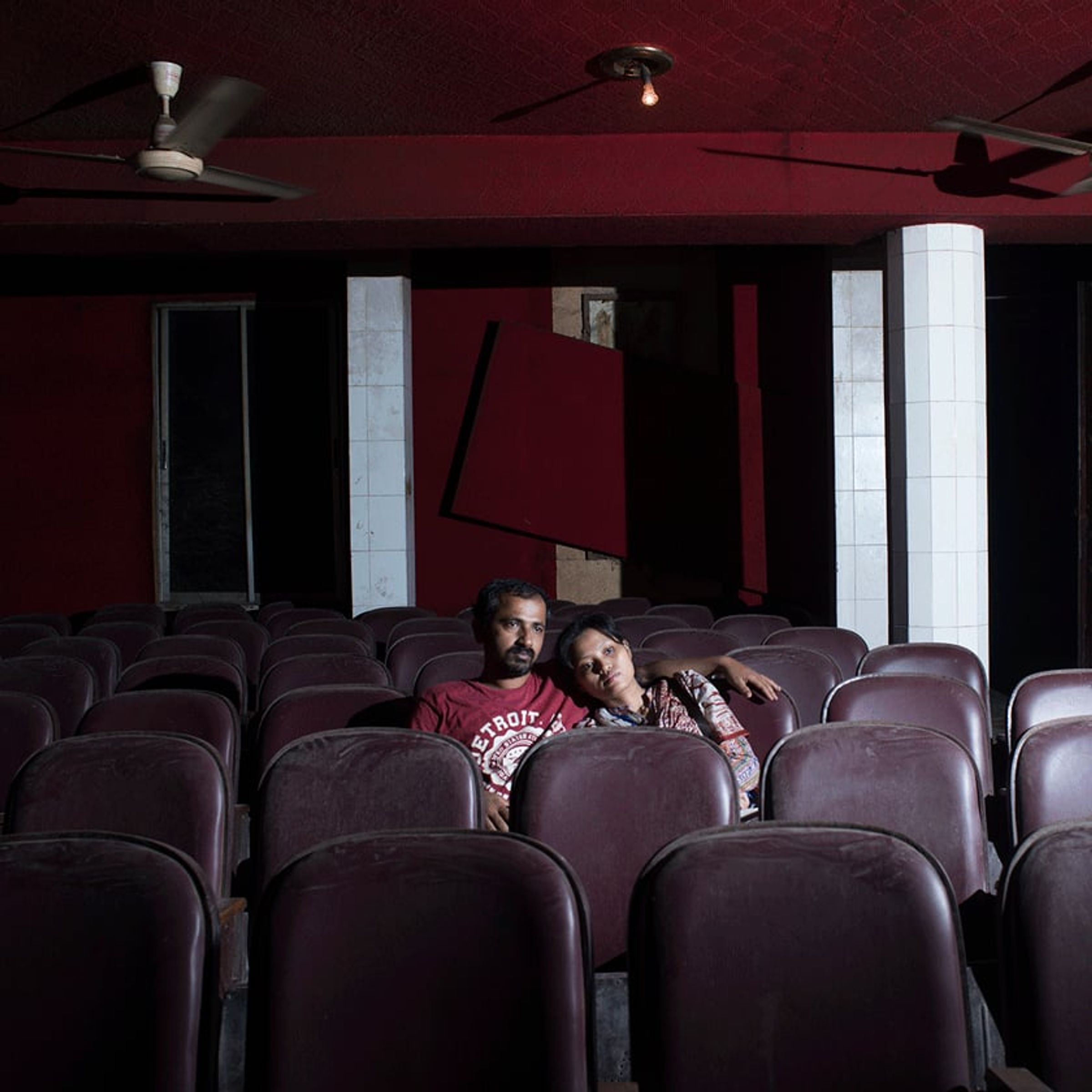 Couple sits close together in an otherwise empty cinema with rows of maroon seats.
