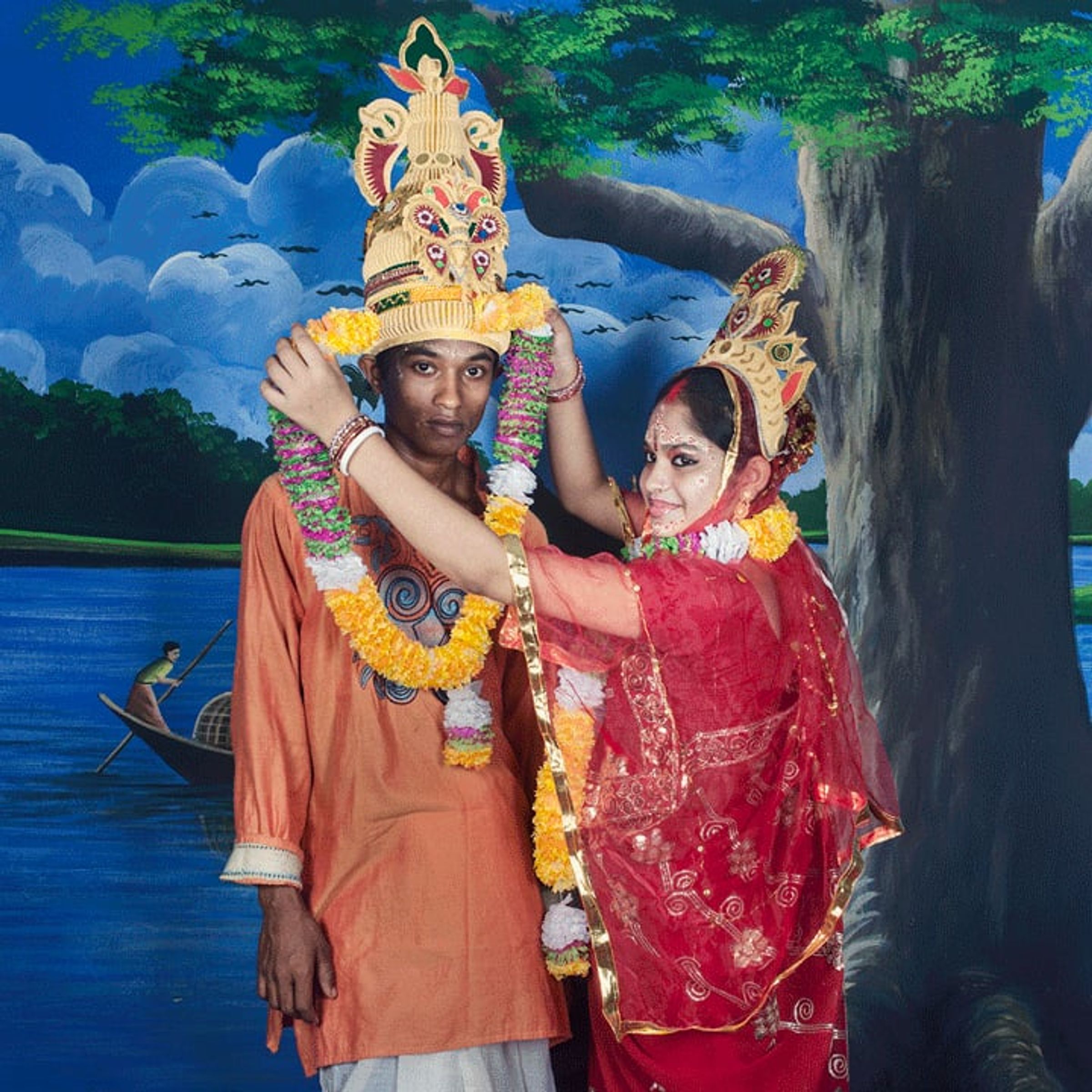 Couple in ornate ceremonial costumes embraces before a painted lakeside backdrop.