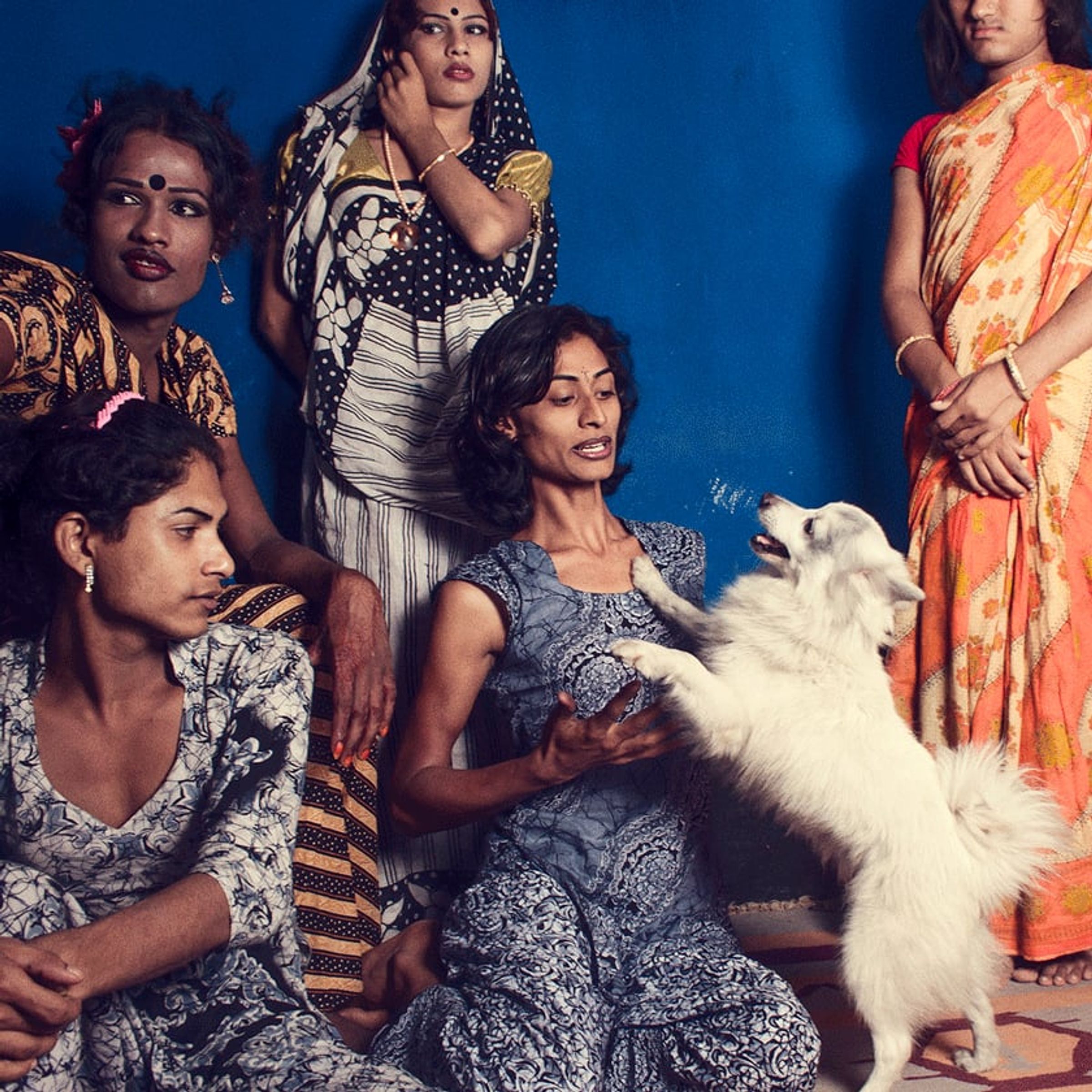 Group of brightly dressed people gathers around a seated person holding a white animal against blue.