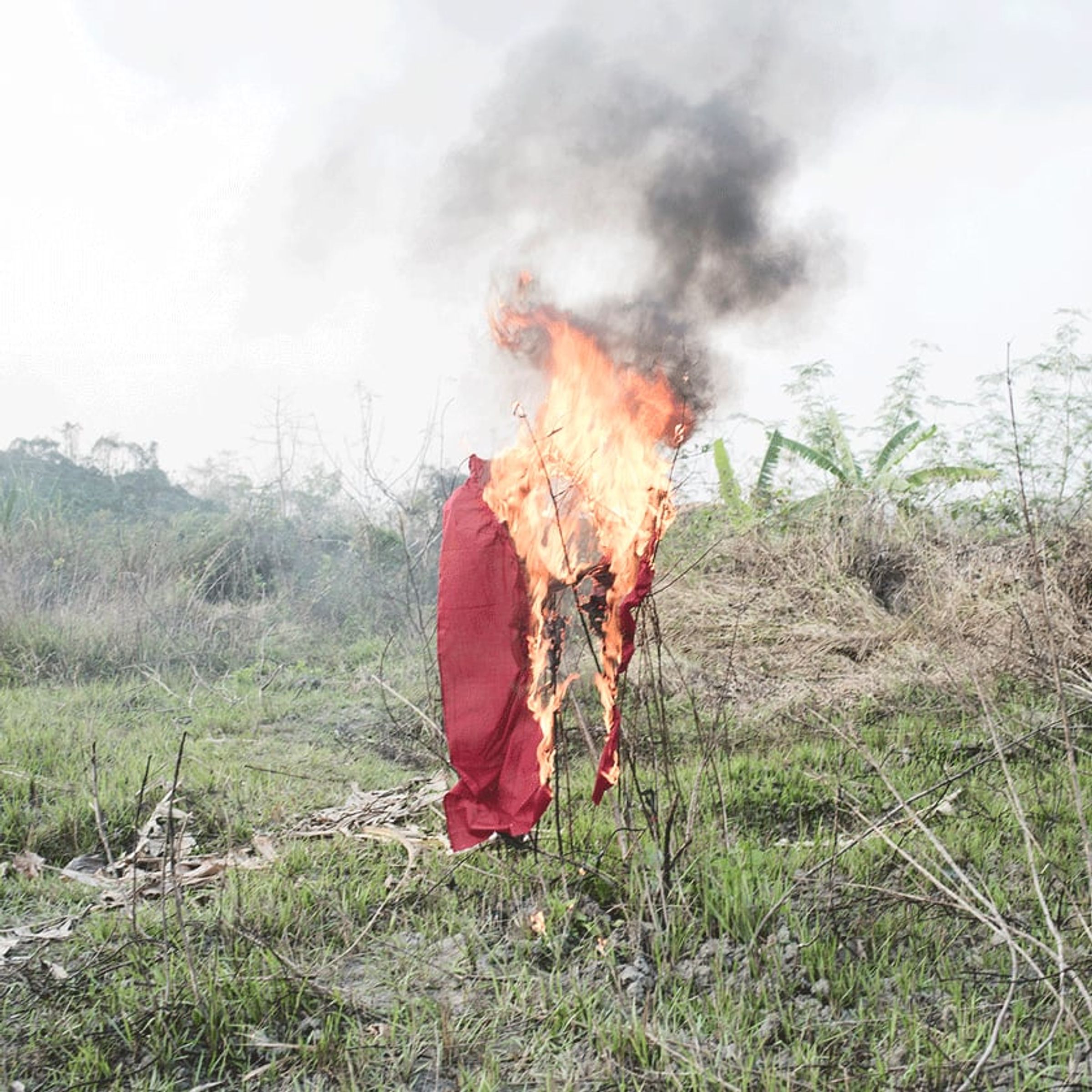 Flames and black smoke consume a red cloth mounted in an open field.
