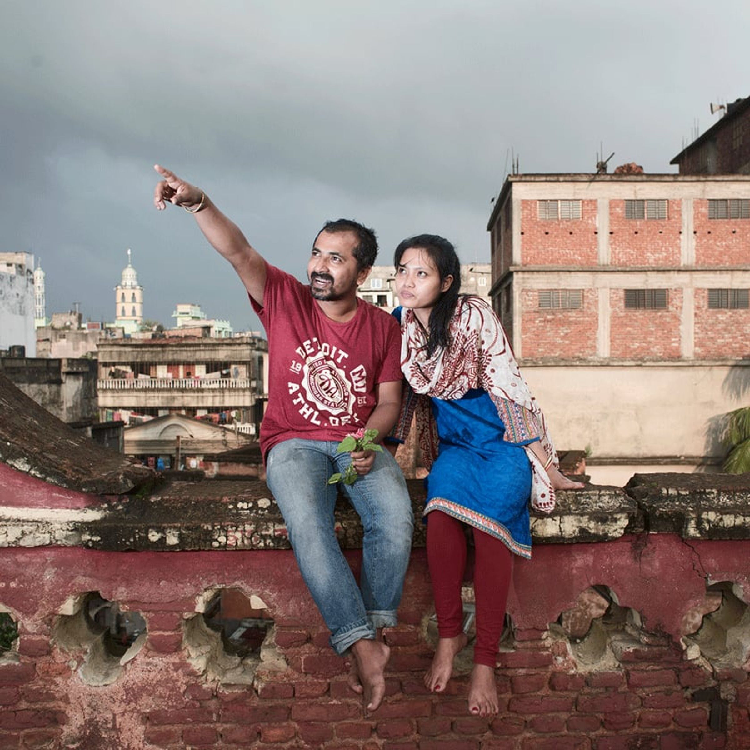 Couple sits on a rooftop ledge, one pointing toward the cloudy skyline.